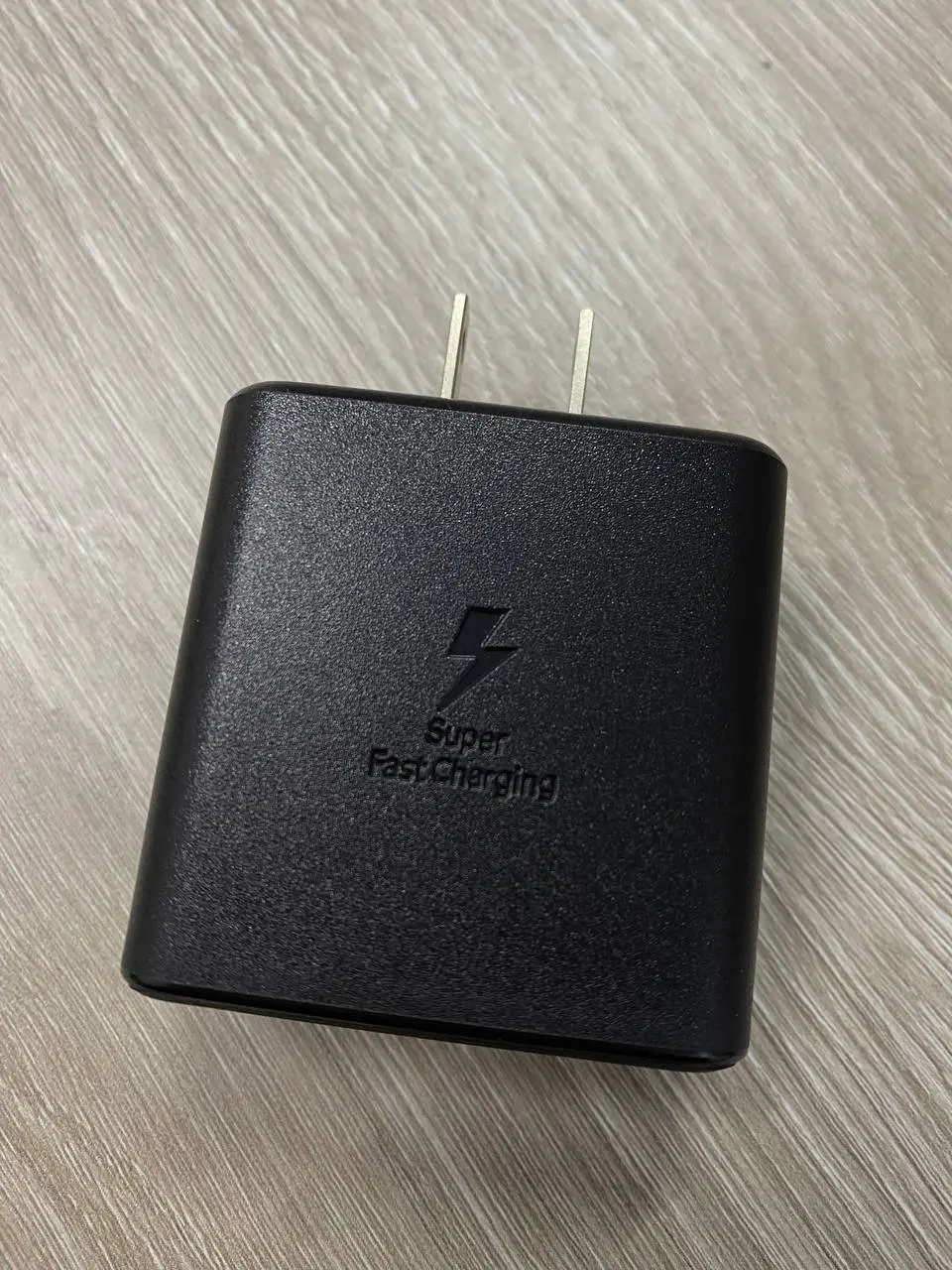 Cargador de pared Samsung negro con textura granulada, logo de Super Fast Charging y dos pines metálicos sobre superficie de madera.