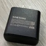 Cargador de pared Samsung negro con puerto USB-C y especificaciones técnicas impresas, colocado sobre superficie de madera.