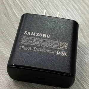 Cargador de pared Samsung negro con puerto USB-C y especificaciones técnicas impresas, colocado sobre superficie de madera.