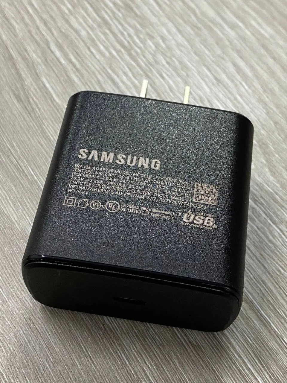 Cargador de pared Samsung negro con puerto USB-C y especificaciones técnicas impresas, colocado sobre superficie de madera.