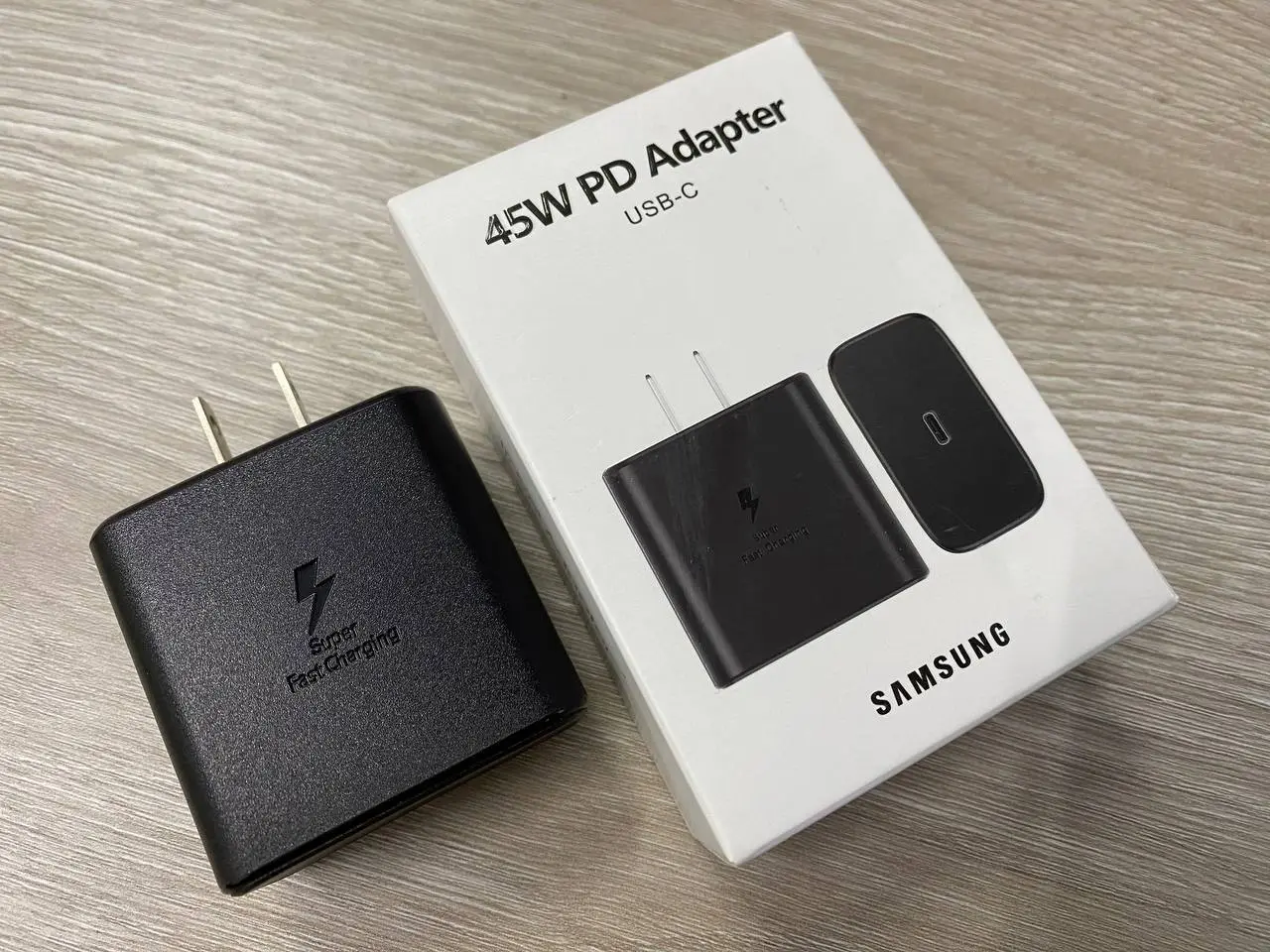 Cargador rápido Samsung negro de 45W con puerto USB-C junto a su caja de empaque blanca sobre superficie de madera.
