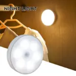Lámpara nocturna LED circular blanca con sensor de movimiento, instalada en la pared de un armario iluminando ropa colgada.