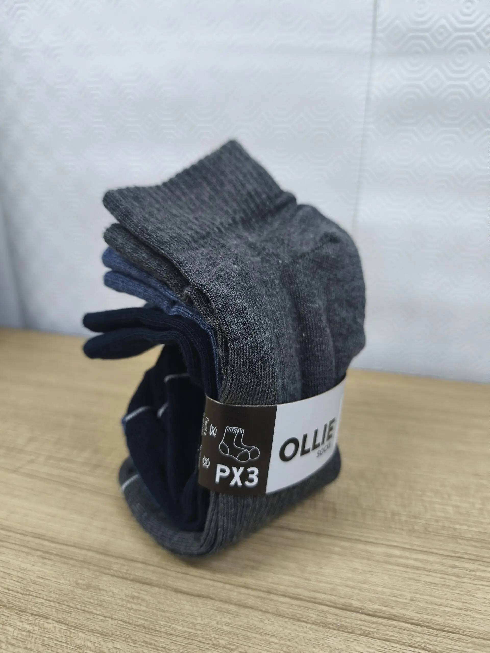 Pack por tres medias tobilleras en tonos gris, azul y marino, dobladas y sujetas con banda de papel de la marca Ollie sobre superficie de madera.