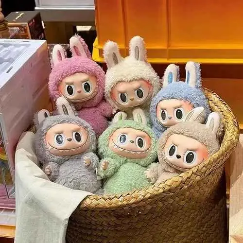 Peluches Labubu con texturas suaves y disfraces de conejo en colores rosa, blanco, azul, gris, verde y crema, dentro de una canasta de mimbre.