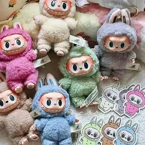 Peluches Labubu de textura suave en colores rosa, verde, azul y crema junto a stickers decorativos sobre una superficie blanca esponjosa.