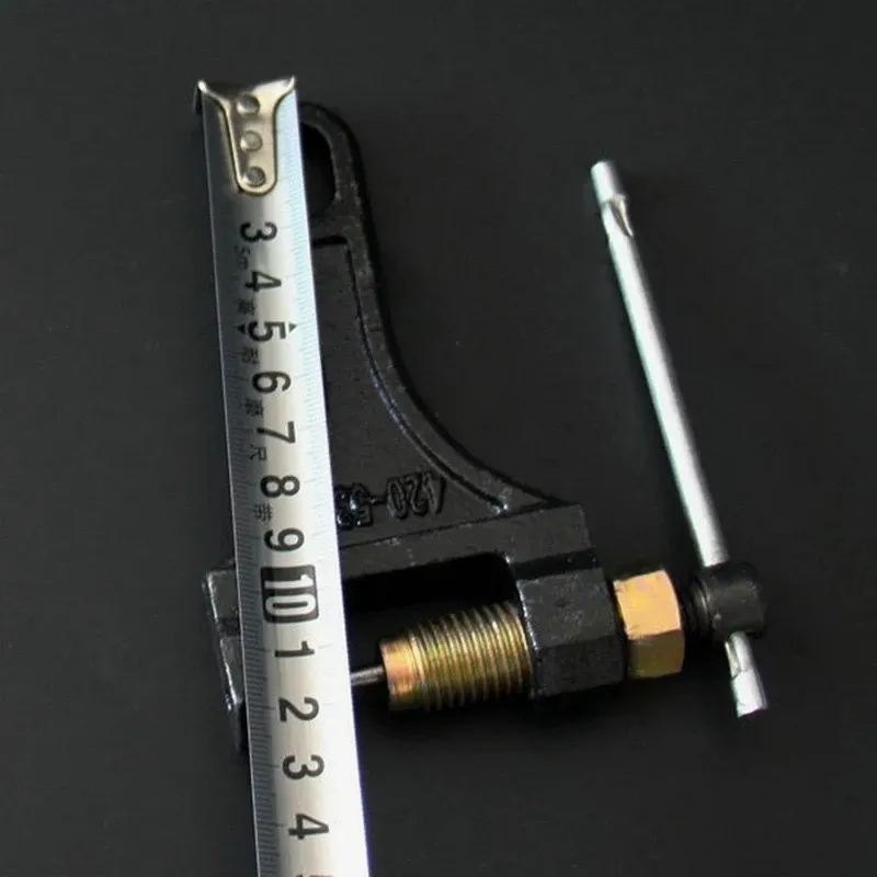 Despinador de cadena para moto de metal negro con tornillo dorado y manivela, junto a una cinta métrica para referencia de dimensiones.