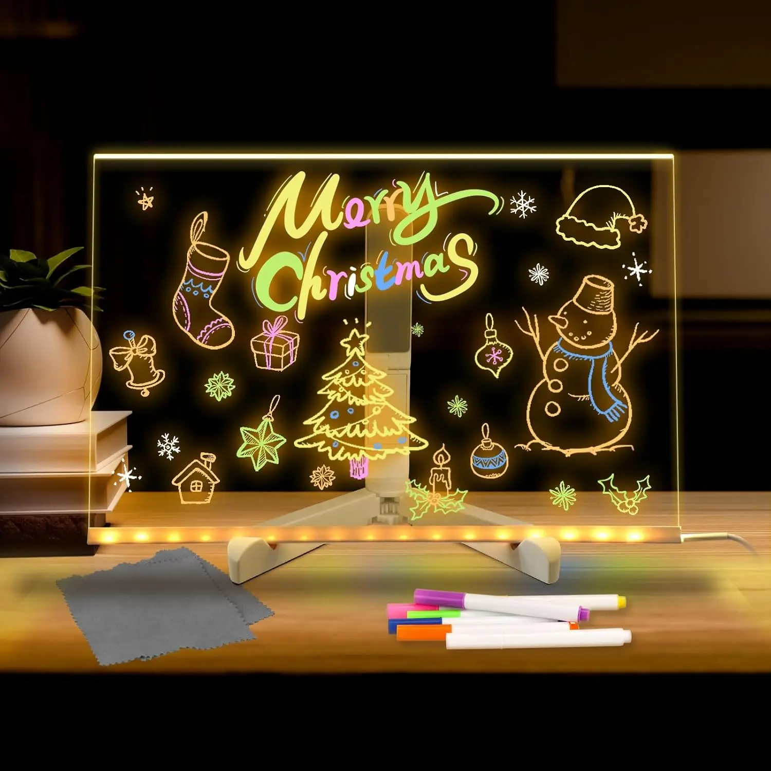 Tablero LED de acrílico transparente con base, incluye marcadores de colores y paño de limpieza, mostrando dibujos navideños iluminados sobre escritorio.