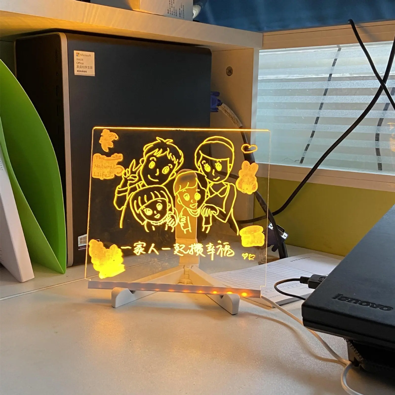 Tablero led de acrílico transparente con base blanca, iluminado en tono naranja con un dibujo familiar grabado y la frase en español sobre un escritorio.