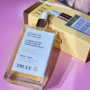 Aceite corporal y para vello púbico Truly Cooka Oil en envase rectangular de vidrio con gotero, acompañado de su caja amarilla sobre superficie rosa.