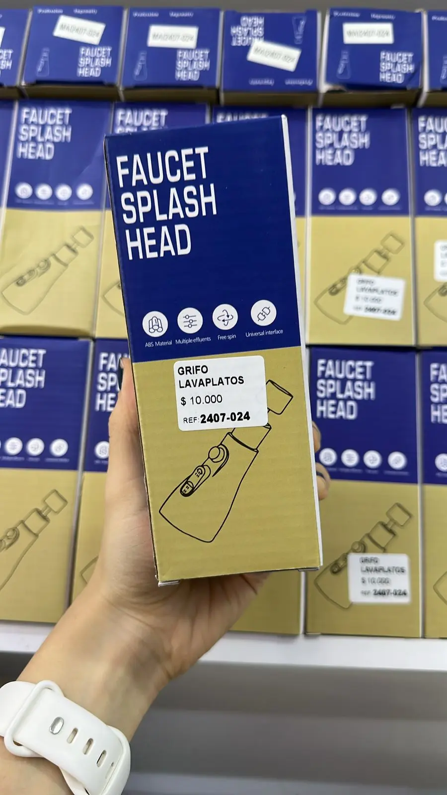 Mano sosteniendo caja marrón y azul de cabezal para grifo de lavaplatos frente a repisa con más unidades idénticas.