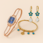 Set de joyería con reloj oro rosa de esfera azul, pulsera dorada con dijes azules y pendientes de flores azules con cristales en gota.