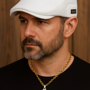 Hombre barbado usando boina plana blanca microperforada con placa lateral oscura y collar de cruz dorada frente a pared de madera.
