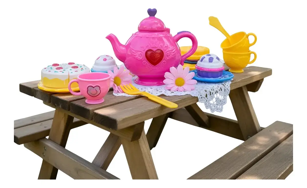 Set de té infantil de juguete en plástico rosa y amarillo, dispuesto sobre una mesa de picnic de madera al aire libre con tetera, tazas, pastel y accesorios.
