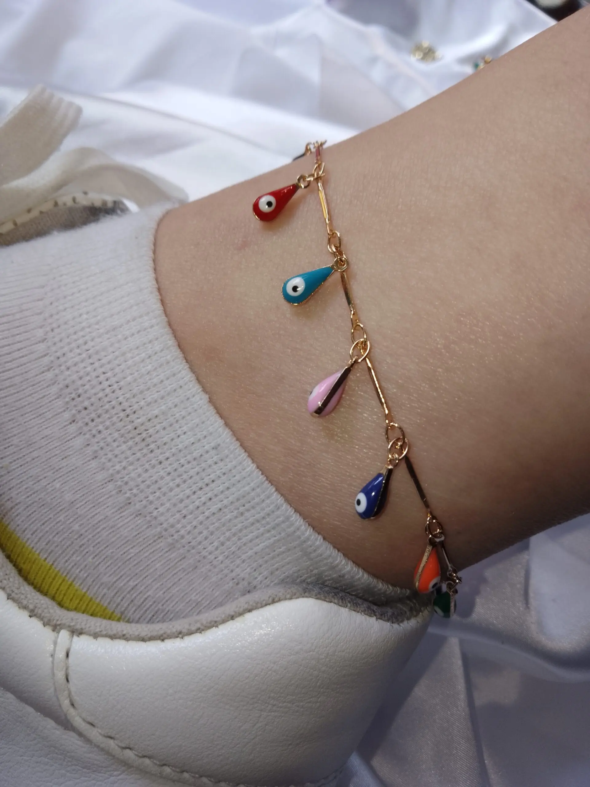 Tobillera dorada con dijes colgantes en forma de gota multicolor y diseño de ojo turco, puesta sobre el tobillo junto a media blanca y zapato.