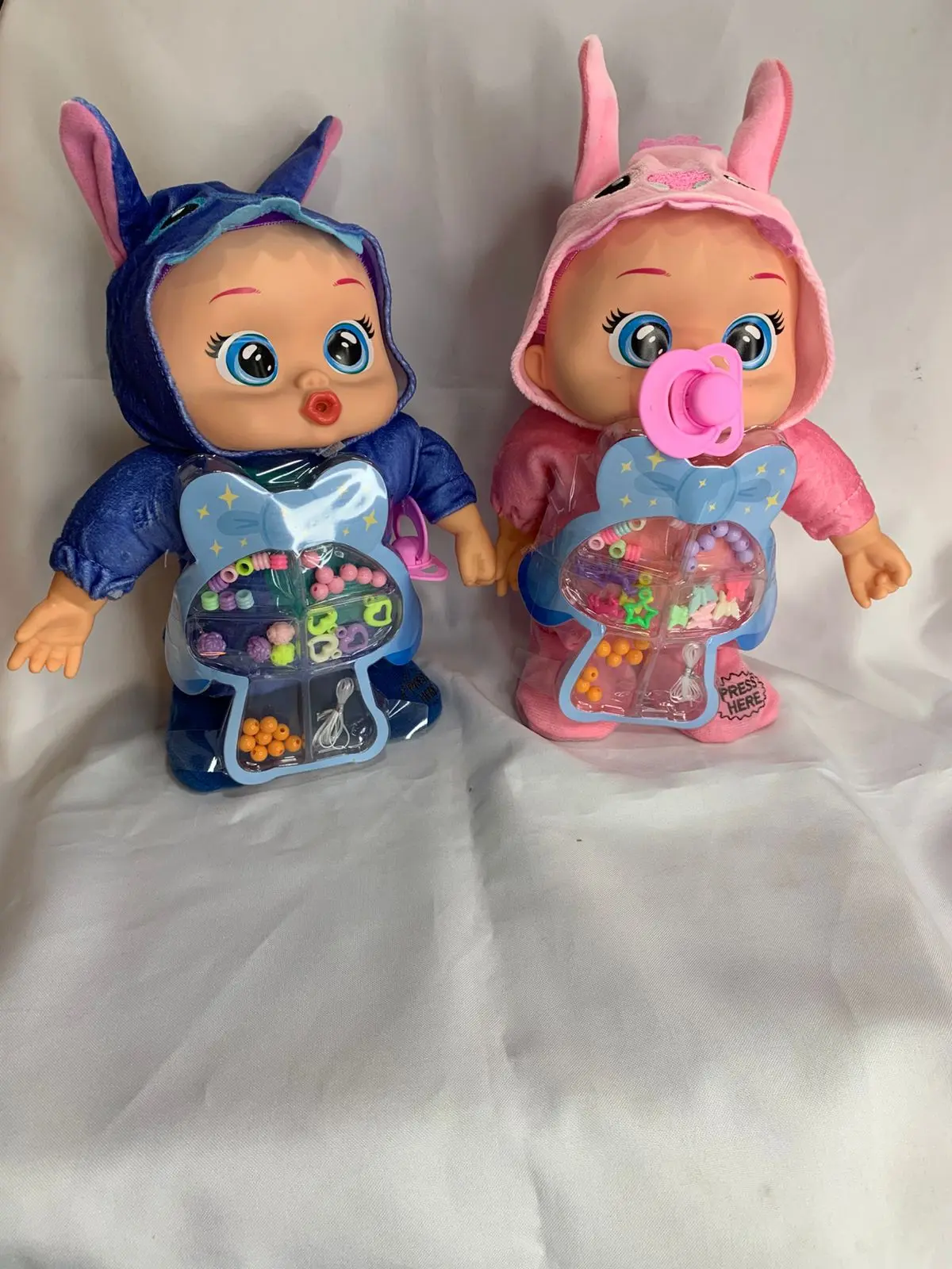 Dos muñecos bebé llorones de peluche, uno azul de Stitch y otro rosa, con capotas de orejas largas y estuches transparentes llenos de cuentas de colores.