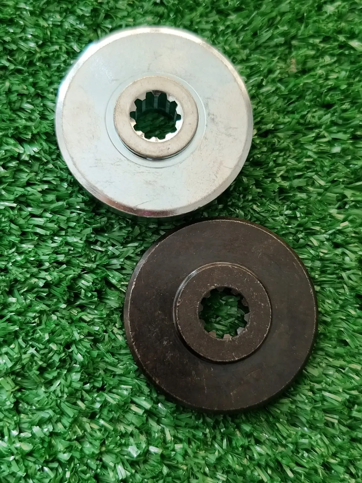 Dos placas de transmisión para guadaña Husqvarna 143RII, una plateada brillante y otra oscura, ambas con centro estriado sobre superficie verde.