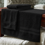 Toalla de baño negra de algodón con textura suave, doblada sobre una mesa de madera junto a toallas blancas.