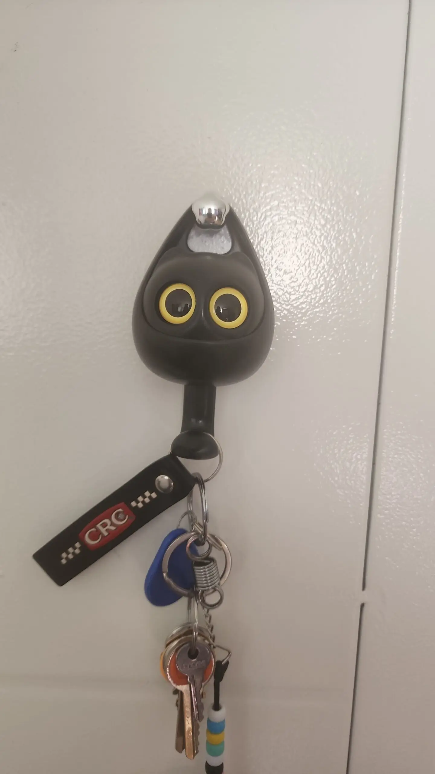 Porta llaves adhesivo con diseño de cabeza de gato negro y ojos amarillos, colgado en una puerta blanca con un manojo de llaves metálicas.