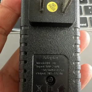 Adaptador de corriente negro con clavijas tipo americano, textura rugosa y salida de 5V 2A, mostrado en vista posterior sostenido por una mano.