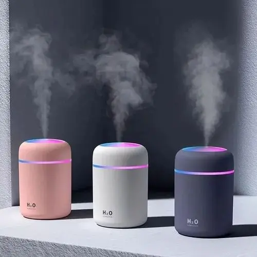Tres humidificadores aromatizadores cilíndricos en rosa, blanco y gris oscuro con luz LED RGB y vapor, sobre superficie blanca.