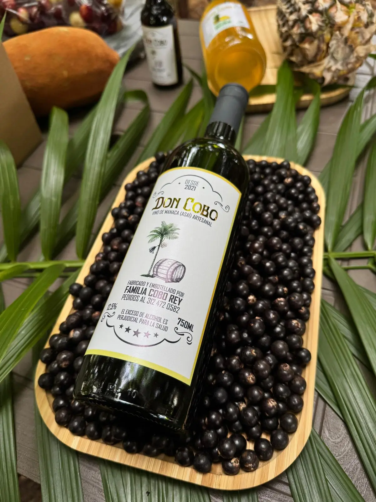 Botella de vino artesanal de asaí sobre bandeja de frutos negros, rodeada de hojas de palma, frutas tropicales y otras botellas en fondo rústico.