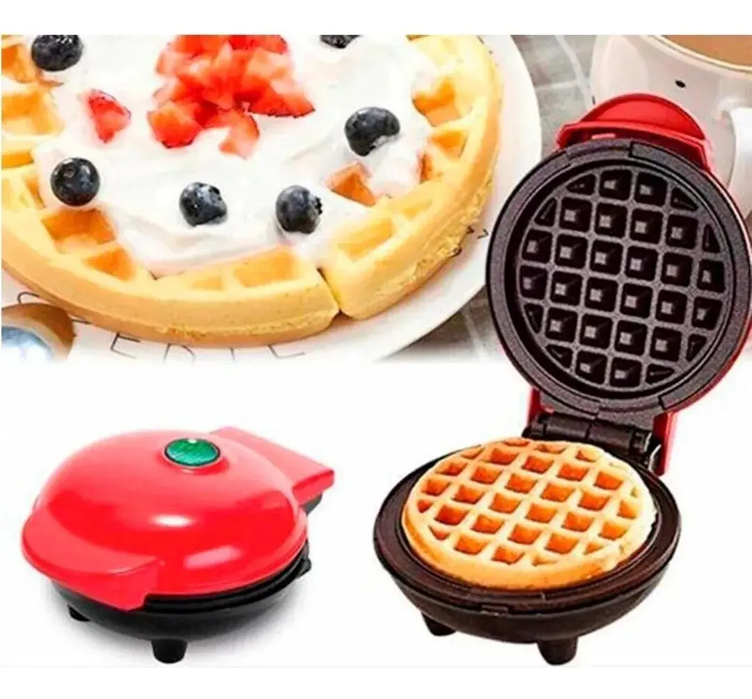 Mini waflera eléctrica roja compacta con superficie antiadherente y indicador luminoso, preparando un waffle redondo junto a un plato servido con crema y frutas.
