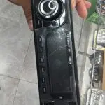 Radio 1 DIN negro con perilla central cromada, botones laterales y pantalla digital, sostenido por una mano en primer plano.