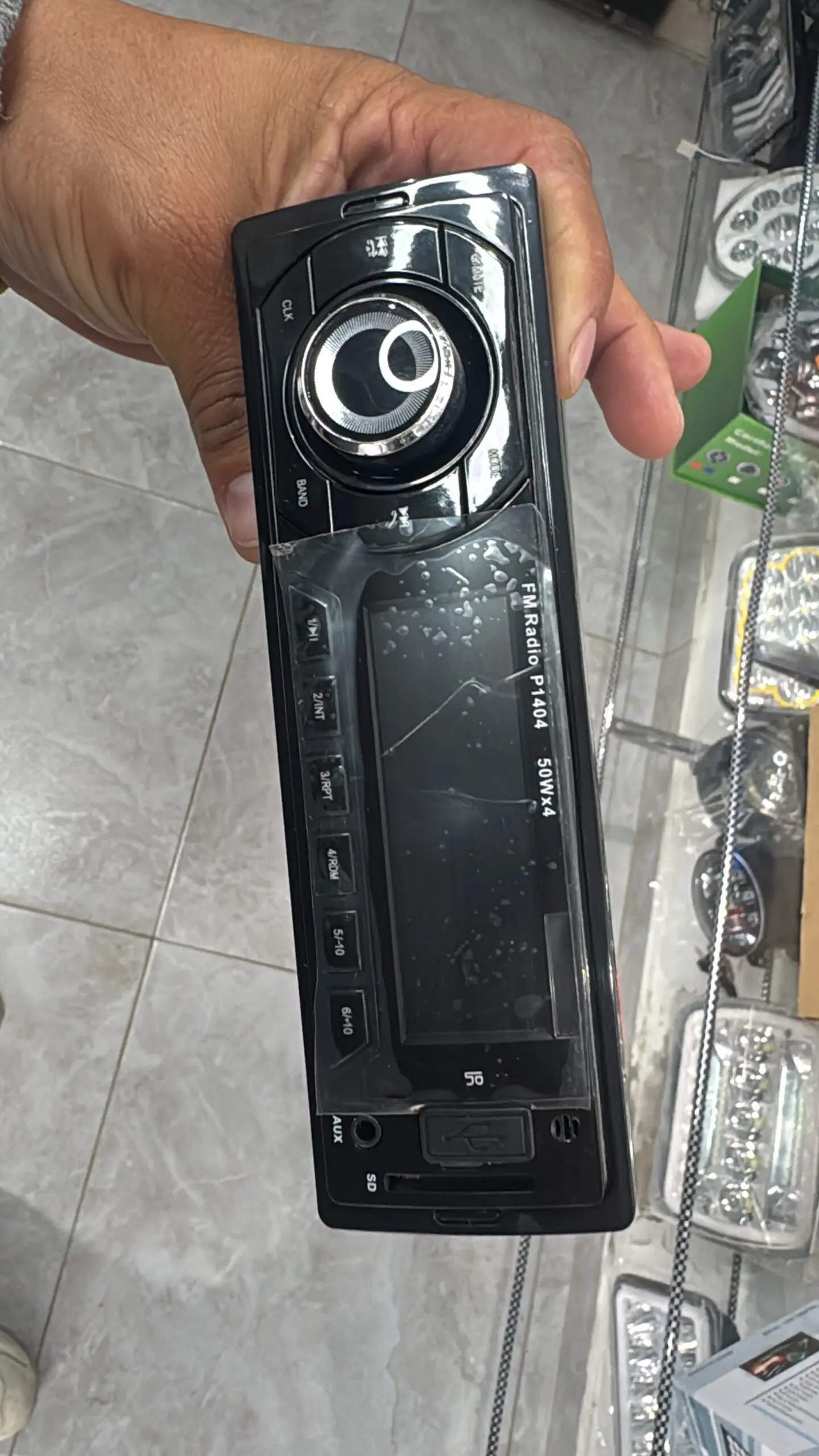 Radio 1 DIN negro con perilla central cromada, botones laterales y pantalla digital, sostenido por una mano en primer plano.
