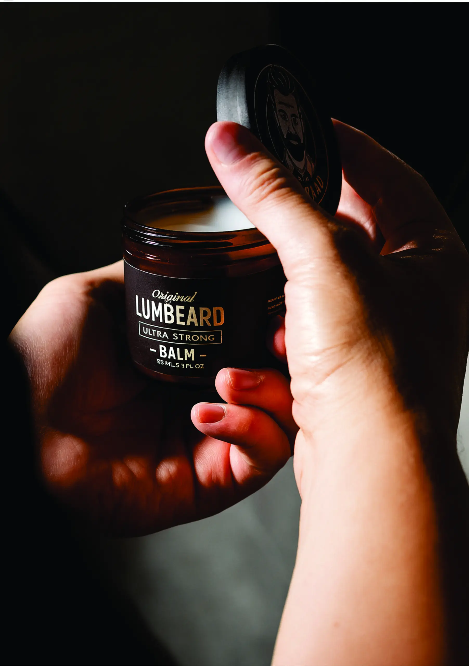 Manos abriendo tarro marrón de bálsamo para barba LUMBEARD ULTRA STRONG con etiqueta negra y texto dorado sobre fondo oscuro.
