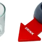 Copa de vidrio transparente y ruleta plástica gris con flecha roja rotulada DRINK para juegos de fiesta.