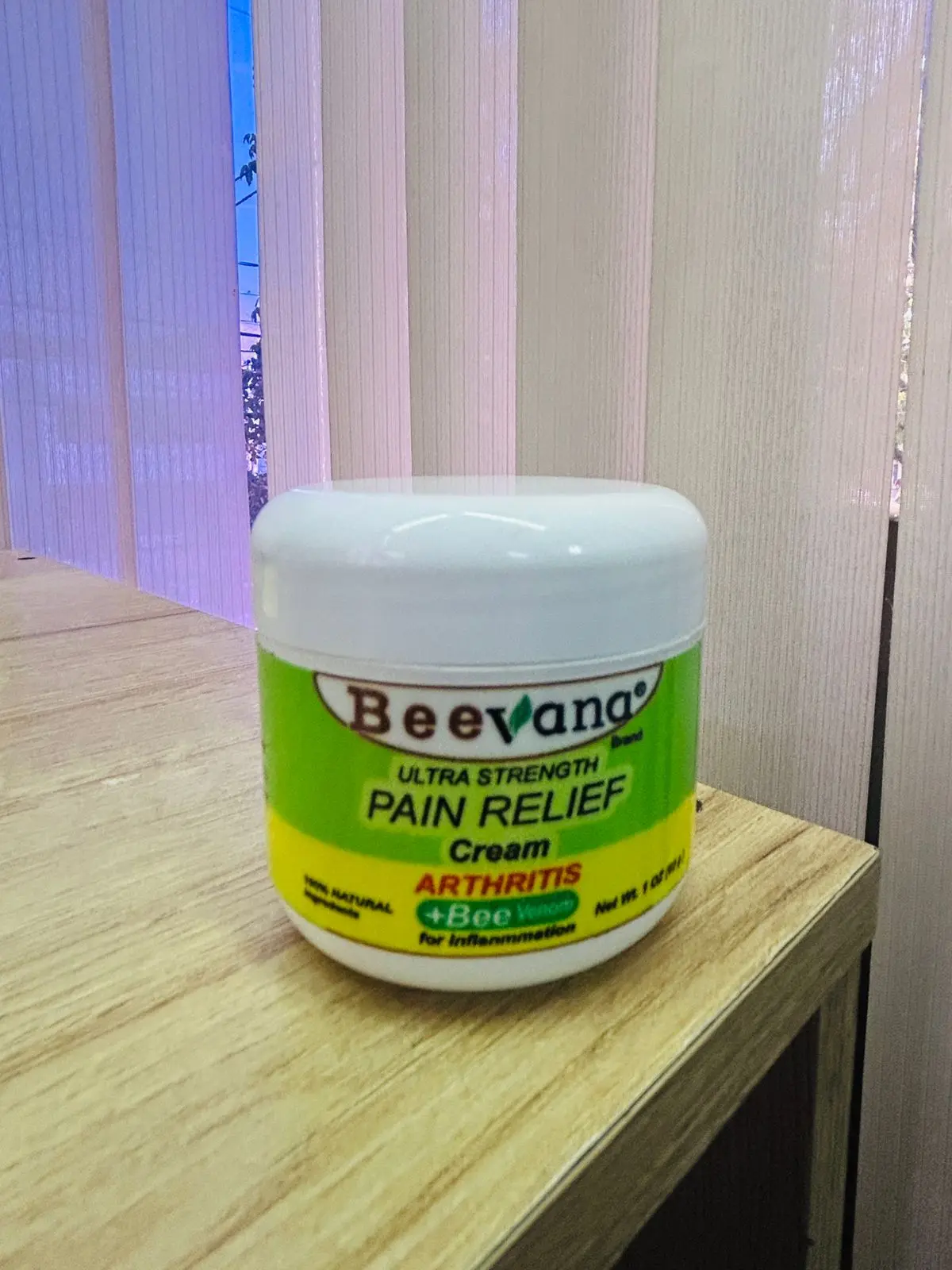 Tarro de crema Beevana para alivio de artritis e inflamación, presentación de 1 oz, sobre superficie de madera con fondo de persianas verticales.