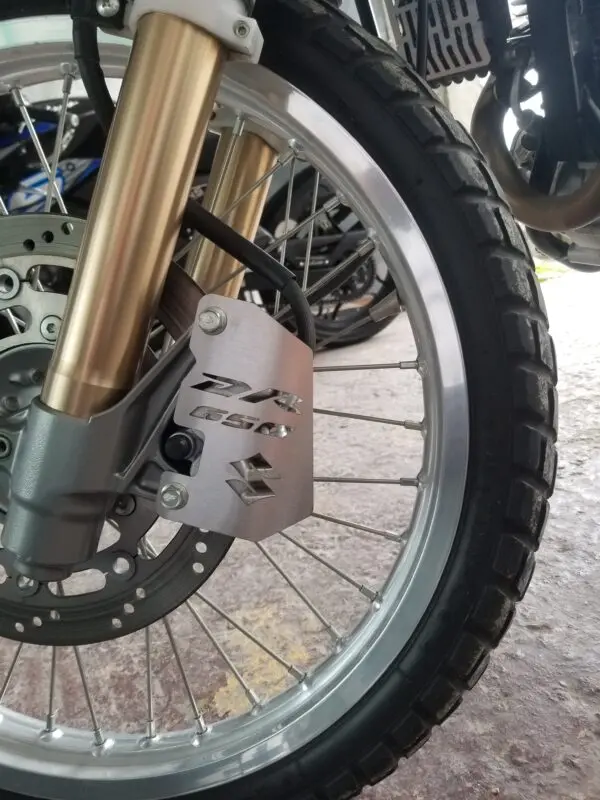 Protector de cáliper de freno en acero inoxidable con grabado DR 650 y Suzuki instalado en la rueda delantera de una motocicleta.