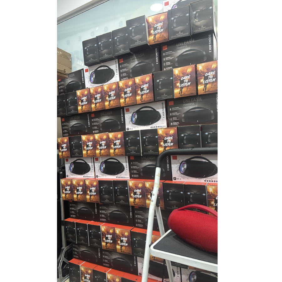 Altavoz Bluetooth JBL Boombox Mini color rojo sobre una escalera blanca, con múltiples cajas de empaque apiladas al fondo.