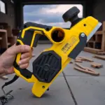 Cepillo eléctrico amarillo y negro de 700W con empuñadura ergonómica y perilla de ajuste, sostenido en mano sobre suelo de concreto con virutas de madera.
