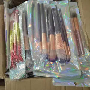 Caja de cartón con varios sets de brochas de maquillaje empacadas en bolsas holográficas, incluyendo mangos metálicos tipo cola de sirena y pinceles con cerdas suaves.