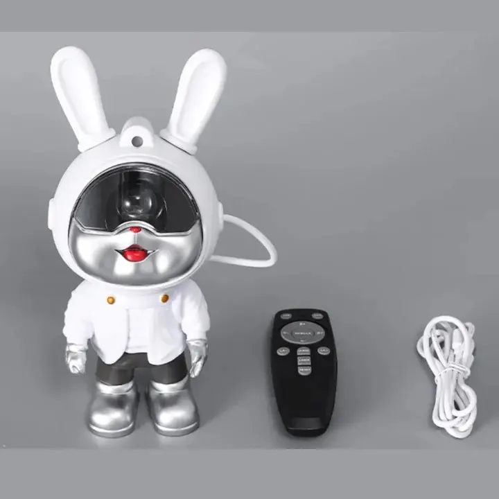 Proyector de galaxias con forma de conejo astronauta blanco, incluye control remoto negro y cable de carga, sobre fondo gris neutro.