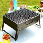 Parrilla BBQ portátil de metal negro con rejilla y patas plegables, dispuesta sobre una mesa blanca en un entorno de picnic al aire libre.