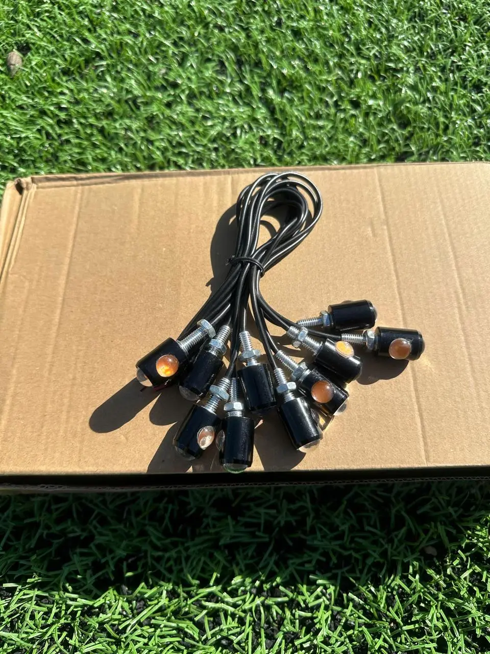 Direccionales LED ojos de águila amarillos con carcasa negra y cables, dispuestos en abanico sobre una caja de cartón en el césped.