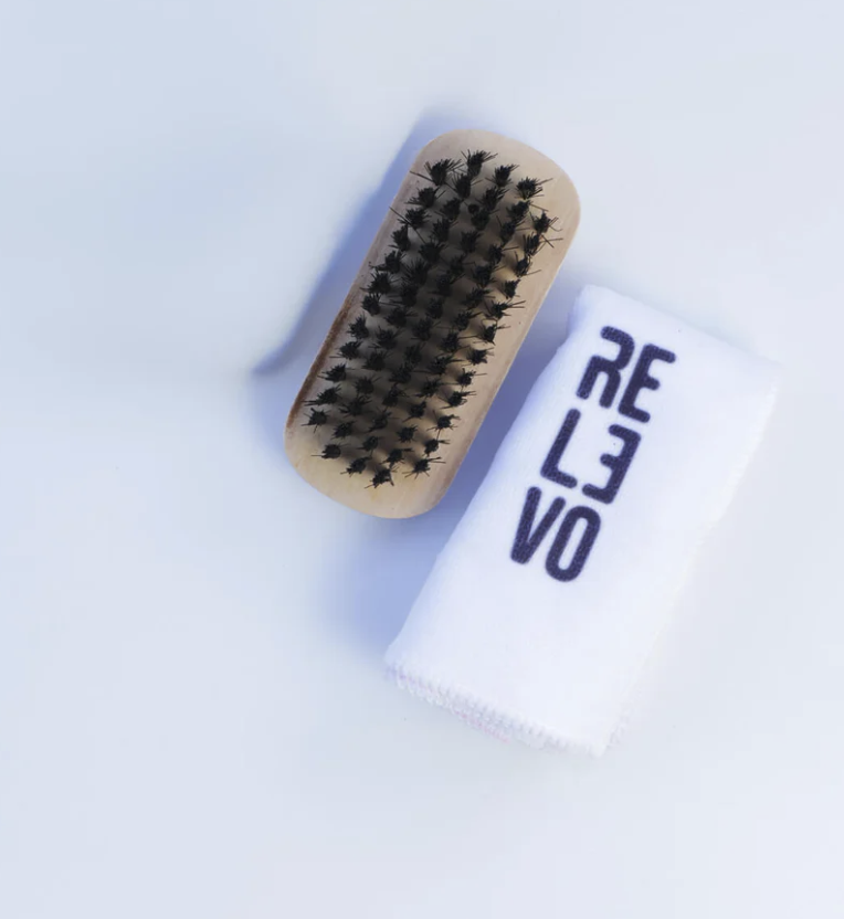 Kit de limpieza para tenis sobre fondo blanco, incluye cepillo de madera con cerdas oscuras y paño blanco con la marca estampada.