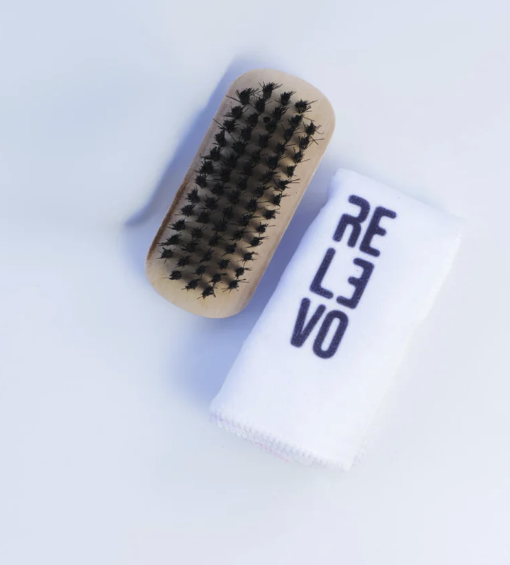 Kit de limpieza para tenis sobre fondo blanco, incluye cepillo de madera con cerdas negras y paño blanco con la marca REVO estampada.