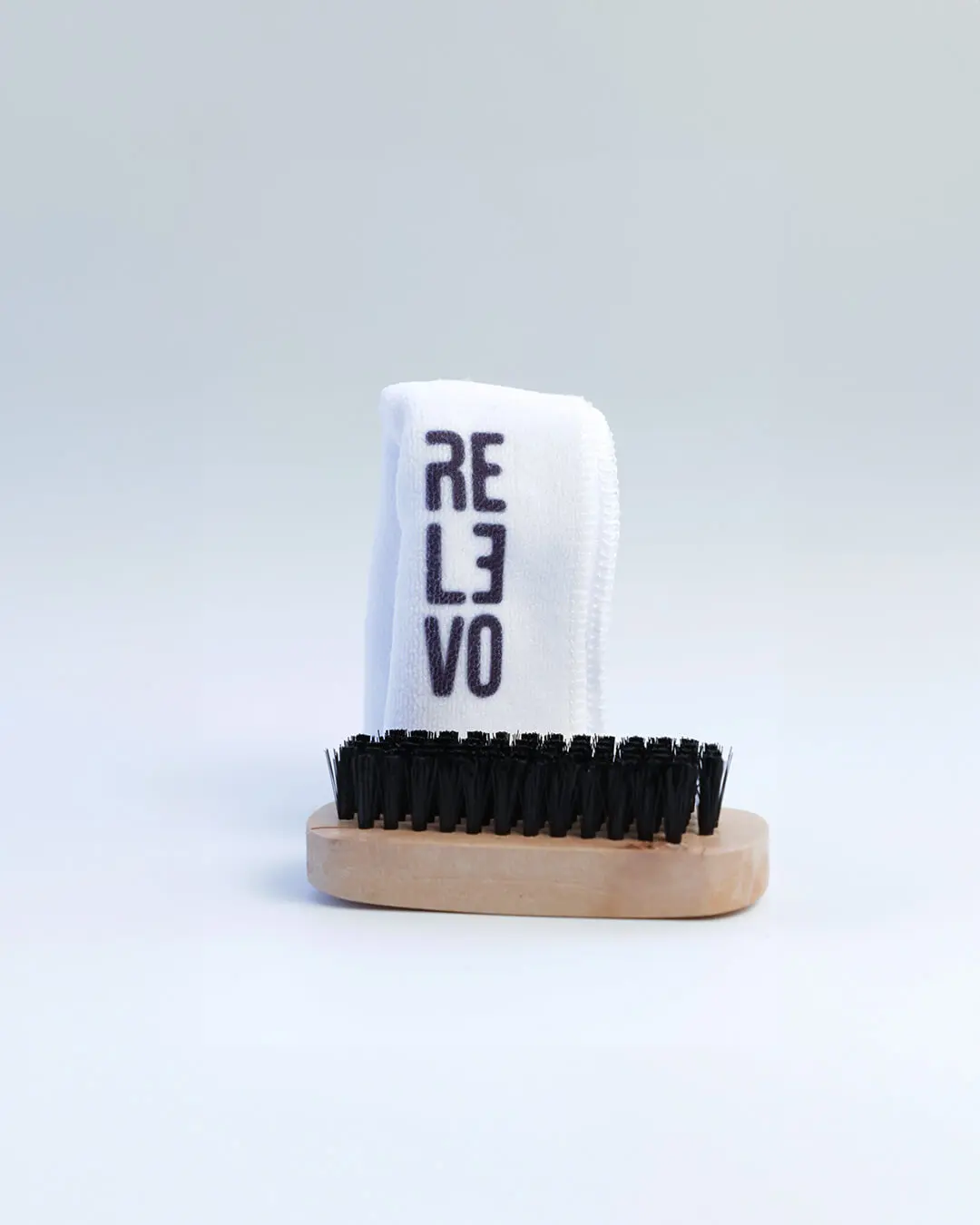 Kit de limpieza para tenis sobre fondo blanco compuesto por cepillo de cerdas negras con base de madera y paño blanco de microfibra con logo REVO.