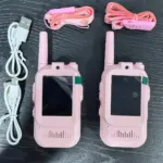 Dos walkie talkies infantiles rosados con cámara y pantalla, acompañados de dos cables USB blancos y dos correas estampadas organizados sobre superficie oscura.