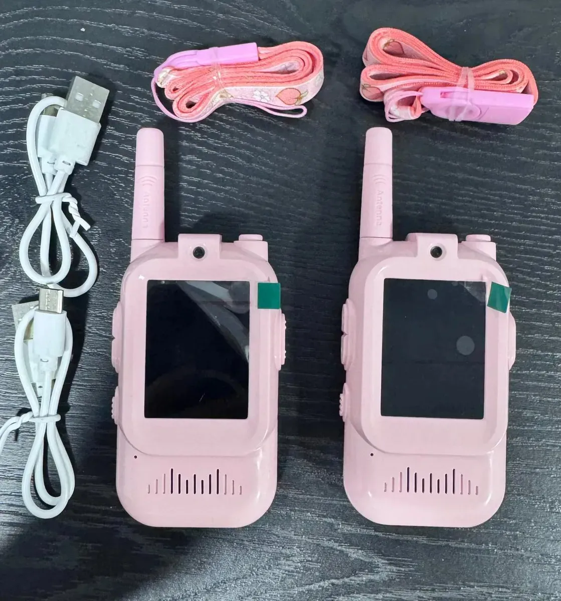 Dos walkie talkies infantiles rosados con cámara y pantalla, acompañados de dos cables USB blancos y dos correas estampadas organizados sobre superficie oscura.