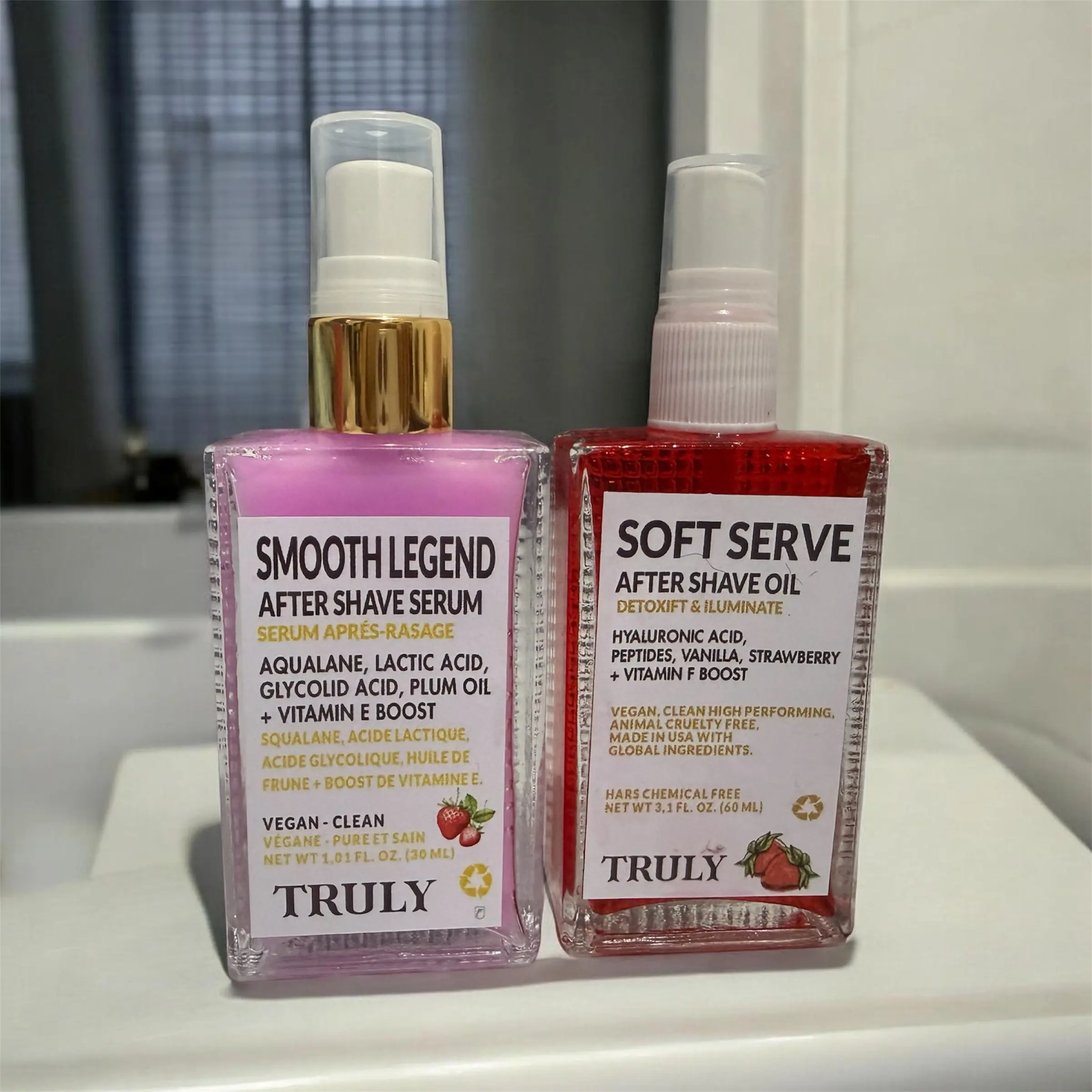 Dúo Truly: sérum Smooth Legend rosa y aceite Soft Serve rojo de 30 ml para después del afeitado, en botellas con dosificador sobre un lavabo.