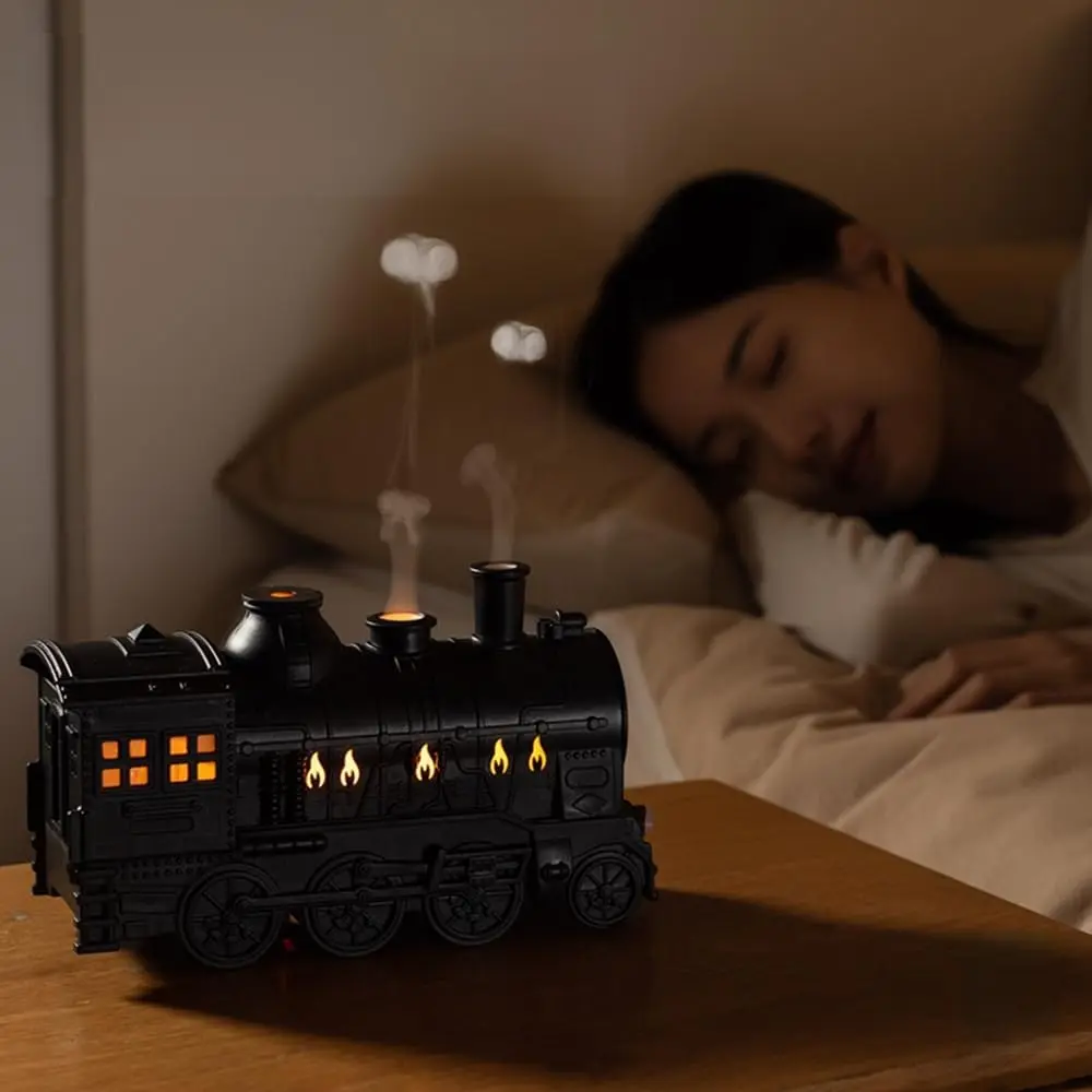Humidificador y difusor de aire negro con diseño de tren antiguo y luz LED cálida tipo fuego, sobre mesa de madera junto a cama con persona durmiendo.