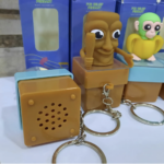 Llavero antiestrés musical marrón con figura animada de moái que abre la boca y emite sonido, mostrado junto a su caja y llavero de metal.