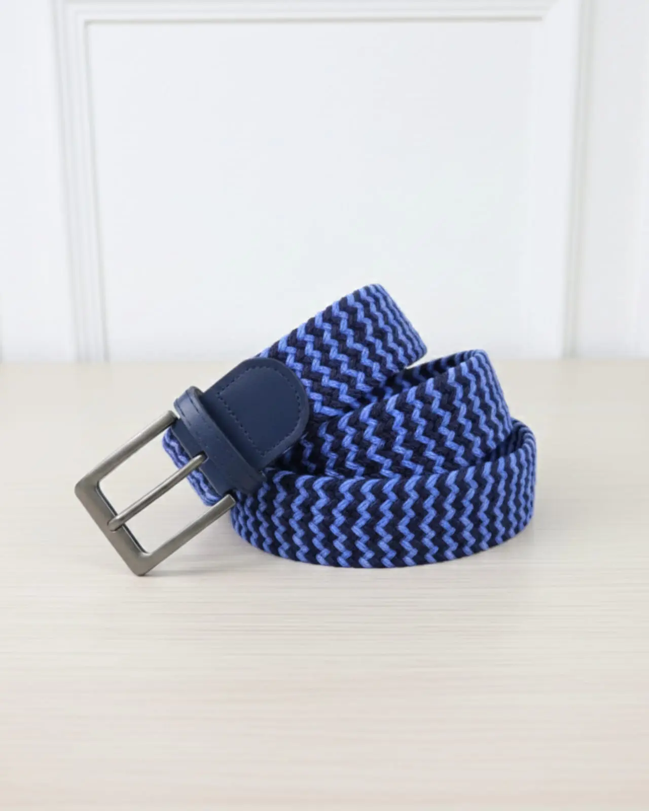 Correa trenzada elástica con patrón zigzag azul y negro, hebilla metálica y detalle en cuero azul sobre superficie clara.