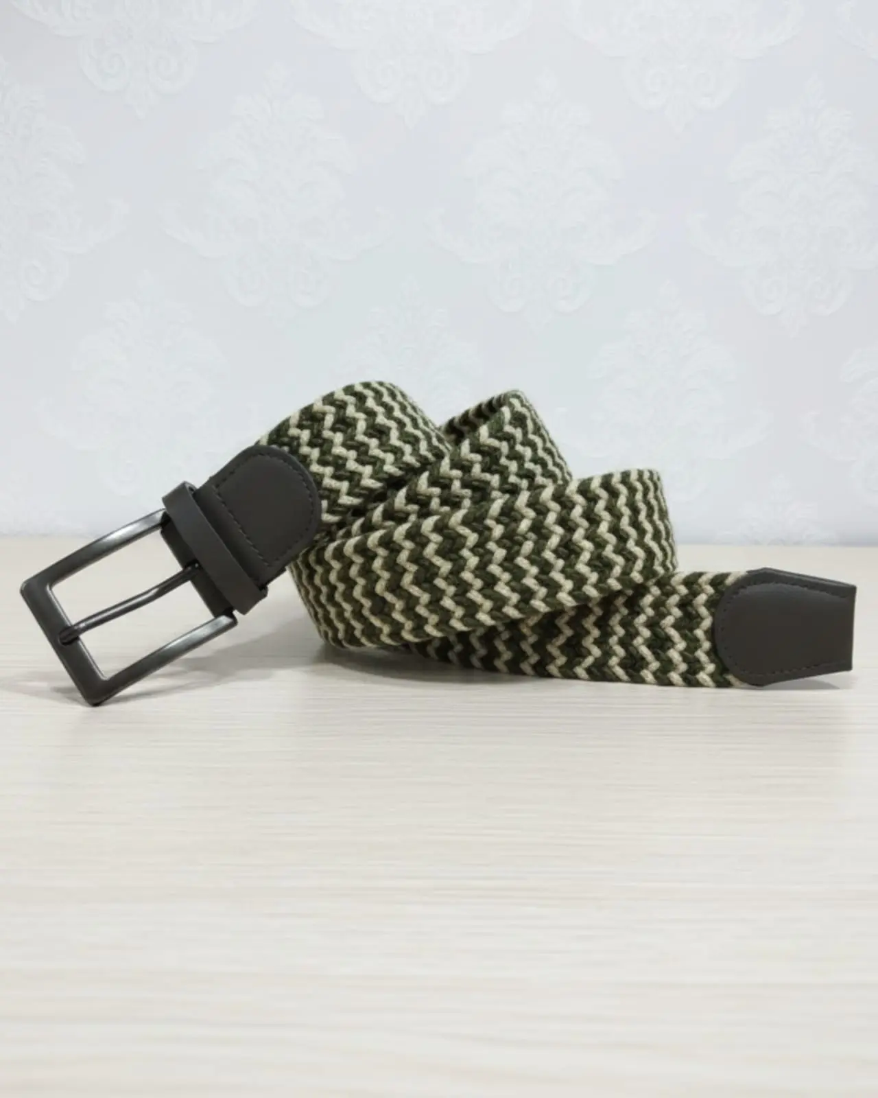 Correa elástica trenzada con patrón zigzag verde oliva y beige, hebilla metálica negra y punta negra, dispuesta de forma curva.