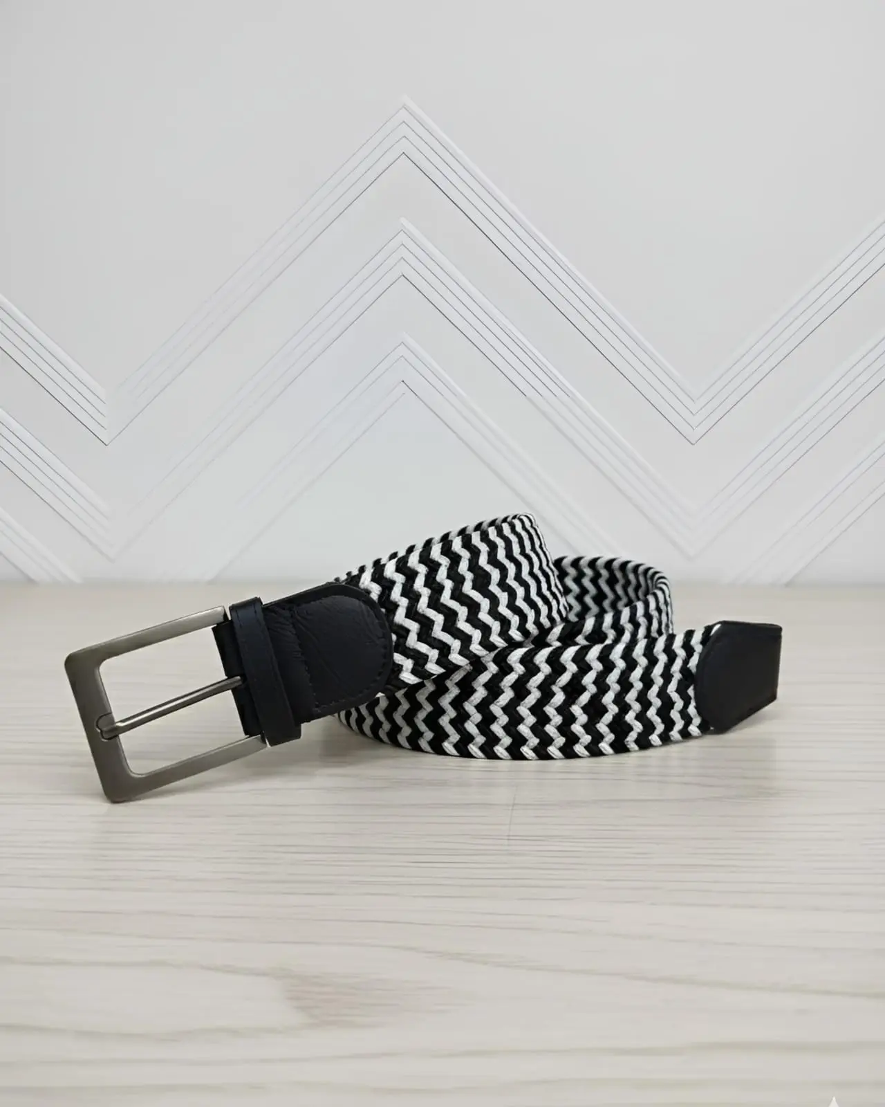 Correa elástica unisex con patrón zigzag blanco y negro, hebilla metálica rectangular gris oscuro y detalles negros sobre superficie de madera.