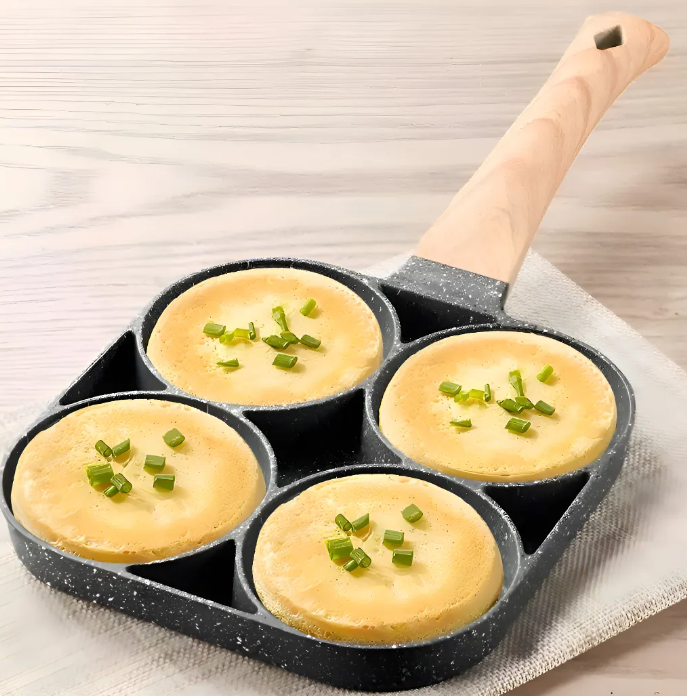 Sartén de aluminio antiadherente gris con cuatro compartimentos circulares, mango de madera y huevos cocidos con cebollín sobre una base de tela.